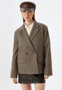 Blazer a quadri marroni con design doppiopetto, rever a scollo, due tasche frontali e bottoni scuri a contrasto, abbinato a una gonna in pelle nera.