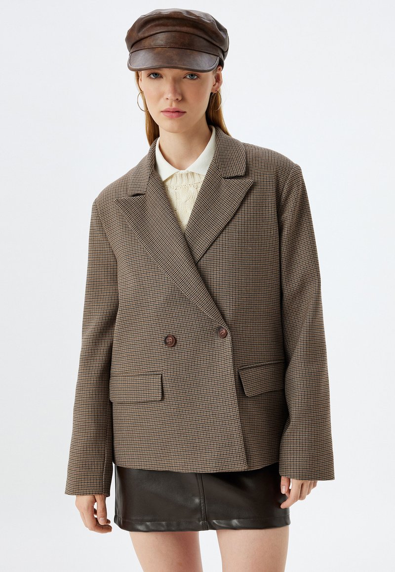 Blazer a quadri marroni con design doppiopetto, rever a scollo, due tasche frontali e bottoni scuri a contrasto, abbinato a una gonna in pelle nera.