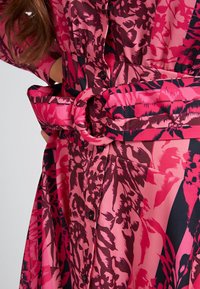 Un vestito a fiori rosa presenta una vita con cintura e fibbia circolare, maniche lunghe e bottoni neri lungo la parte frontale.