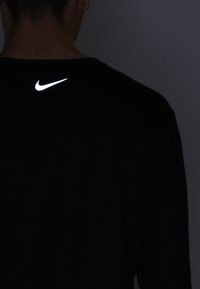 Schwarzes sportliches Shirt mit strukturiertem Muster; mit einem reflektierenden weißen Nike-Logo auf der oberen Rückseite.
