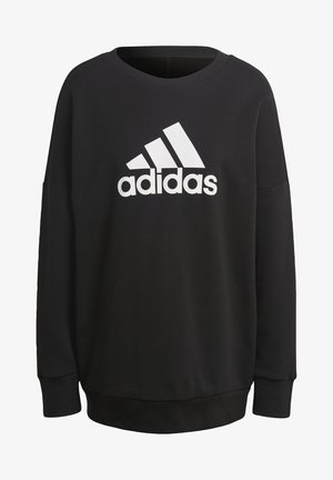 Personne portant un sweat Adidas noir, un pantalon de jogging gris foncé avec des bandes latérales, et des baskets noires à semelles blanches, debout avec un pied en avant.