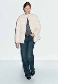 Femme portant une veste en duvet crème sur une chemise grise, un jean large bleu foncé et des chaussures noires, marchant devant un fond clair.