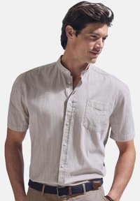 Short-sleeve, light beige button-up shirt with a textured fabric, a chest pocket, and a band collar. Worn with a navy braided belt.