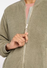 Maslinasto zelena flis bomber jakna s patentnim zatvaračem, rebrastim manžetama i glatkom unutarnjom podstavom. Ima mekanu, teksturiranu vanjštinu.