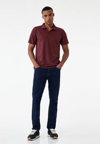 Weinrote Polo-Shirt, kurze Ärmel, Kragen, kombiniert mit dunkelblauen Jeans. Modell trägt schwarze Sneakers mit weißen Schnürsenkeln. Glattes Material, lässiger Stil.