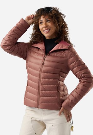 Mujer con chaqueta acolchada rosa polvoriento y pantalones crema, sonriendo y ajustándose las gafas de sol en su cabello rizado, con llaves sujetas en su bolsillo.