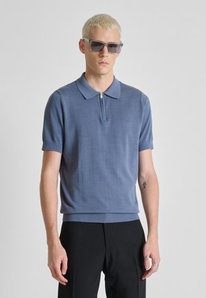 Blonde man met vierkante zonnebril, blauw shirt met korte mouwen en ritskraag, en zwarte broek, staand tegen een effen lichte achtergrond.