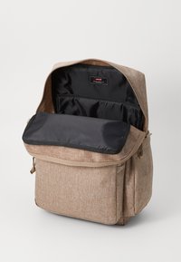Mochila de tela marrón con un exterior texturizado, solapa abierta que revela un bolsillo interior negro y forro, con correas para los hombros y bolsillo frontal.