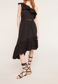 Frau in einem schwarzen einseitig geschnittenen Kleid mit Lochdetails und asymmetrischem Saum, kombiniert mit schwarzen, flachen Schnürriemchenschuhen.