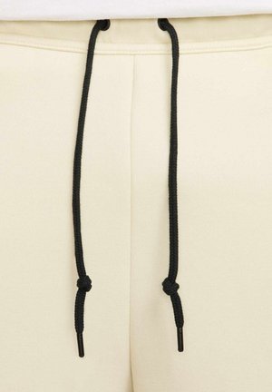 Pantaloni sportivi beige con una texture liscia, dotati di lacci neri con punte in plastica, che mostrano un design semplice e funzionale.