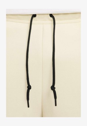 Pantaloni sportivi beige con una texture liscia, dotati di lacci neri con punte in plastica, che mostrano un design semplice e funzionale.