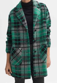 Grün, grau und schwarz karierter Mantel mit großen Revers, vorderen Taschen und schwarzen Knöpfen, getragen über einem schwarzen Rollkragenpullover.