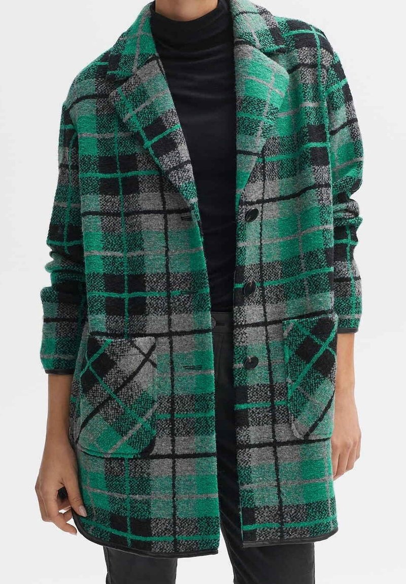 Grün, grau und schwarz karierter Mantel mit großen Revers, vorderen Taschen und schwarzen Knöpfen, getragen über einem schwarzen Rollkragenpullover.