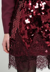 Dłoń o bladej karnacji spoczywająca obok bordowej spódnicy z cekinami i koronką na czarnych rajstopach, na neutralnym tle.