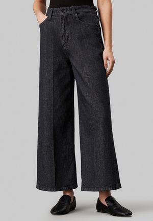 Zwarte denim jeans met wijde pijpen gedragen met zwarte instapschoenen, handen deels in de zakken, staand tegen een effen grijze achtergrond.