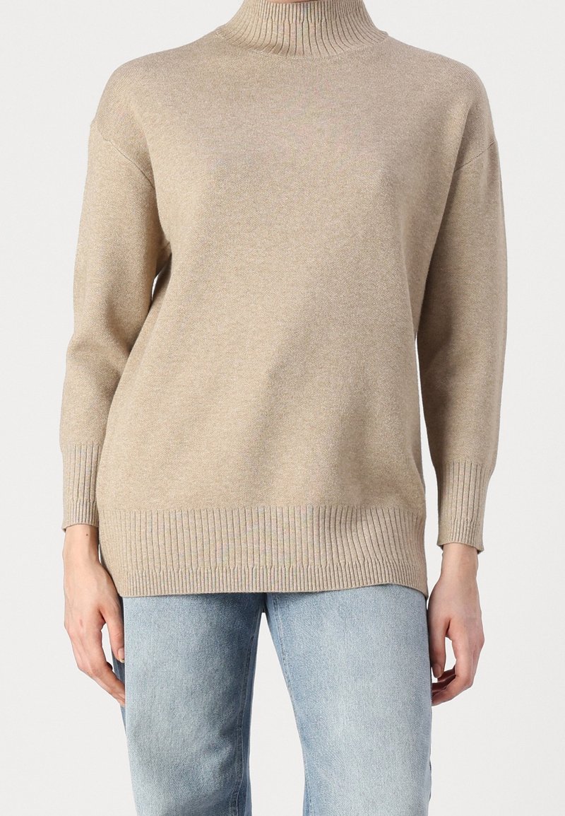 Pull à col roulé côtelé beige avec manches longues associé à un jean bleu clair sur une personne debout.