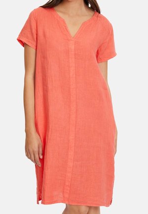 Vrouw draagt een losse koralen linnen jurk tot op de knie met korte mouwen en V-hals, staand tegen een effen achtergrond.