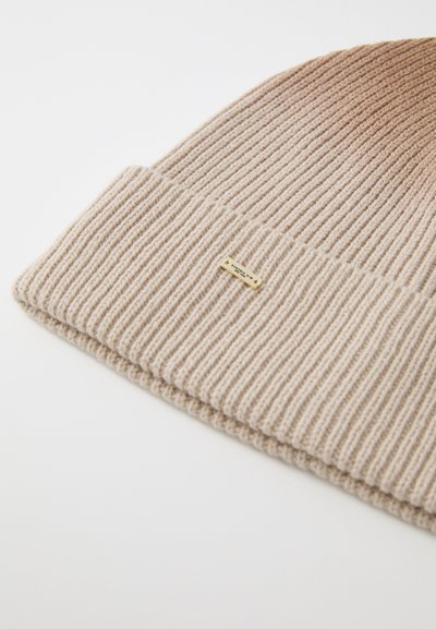 Kootud beež beanie ribatreorigiga, mille esiosal on väike kuldne logo silt ja allpool volt. 
