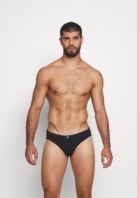 Boxers noirs avec une texture lisse et une coupe basse, arborant un logo discret sur la taille, portés par un mannequin masculin au physique athlétique.