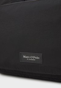 Sac en tissu noir au toucher lisse, fermeture éclair, et une étiquette rectangulaire noire avec du texte blanc, affichant le nom de la marque et les détails de l'établissement.