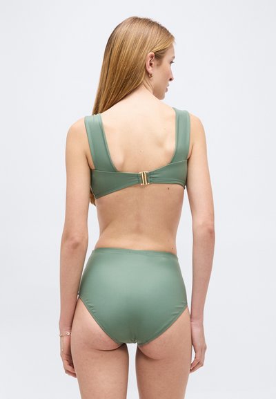 Kvinne med langt blondt hår iført en salviegrønn høytlivs bikiniunderdel og tilpasset bikinitopp, stående mot en ensfarget lys bakgrunn.