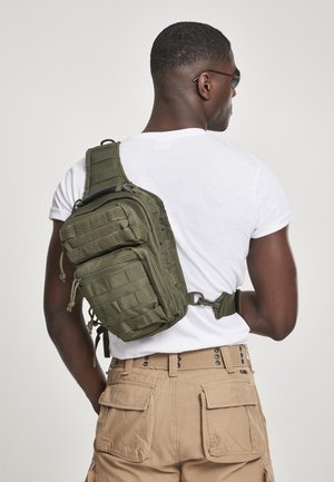 Green tactical sling bag worn across the back, featuring a textured surface, multiple compartments, and adjustable strap.