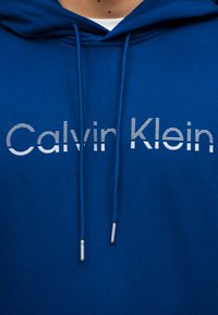 Μπλε φούτερ με κουκούλα, ρυθμιζόμενα κορδόνια και λευκό λογότυπο Calvin Klein στο στήθος.