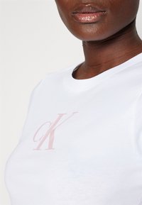 Primer plano de una persona que lleva una camiseta blanca con un logo "cK" de color rosa pálido centrado en el pecho y labios brillantes visibles.