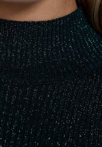 Donkergroen geribbelde gebreide trui met een hoge kraag, met metallic draden die glans en textuur toevoegen. Close-up weergave.