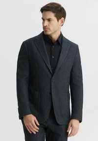 Ciemnoszary blazer wykonany z teksturowanej tkaniny, z klapami w kształcie notcha, dwoma przednimi kieszeniami oraz zapięciem na jeden guzik. Połączony z czarną koszulą.