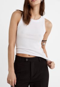 Débardeur blanc à côtes avec un col rond, associé à un pantalon noir taille haute avec une fermeture à bouton et des plis frontaux marqués.