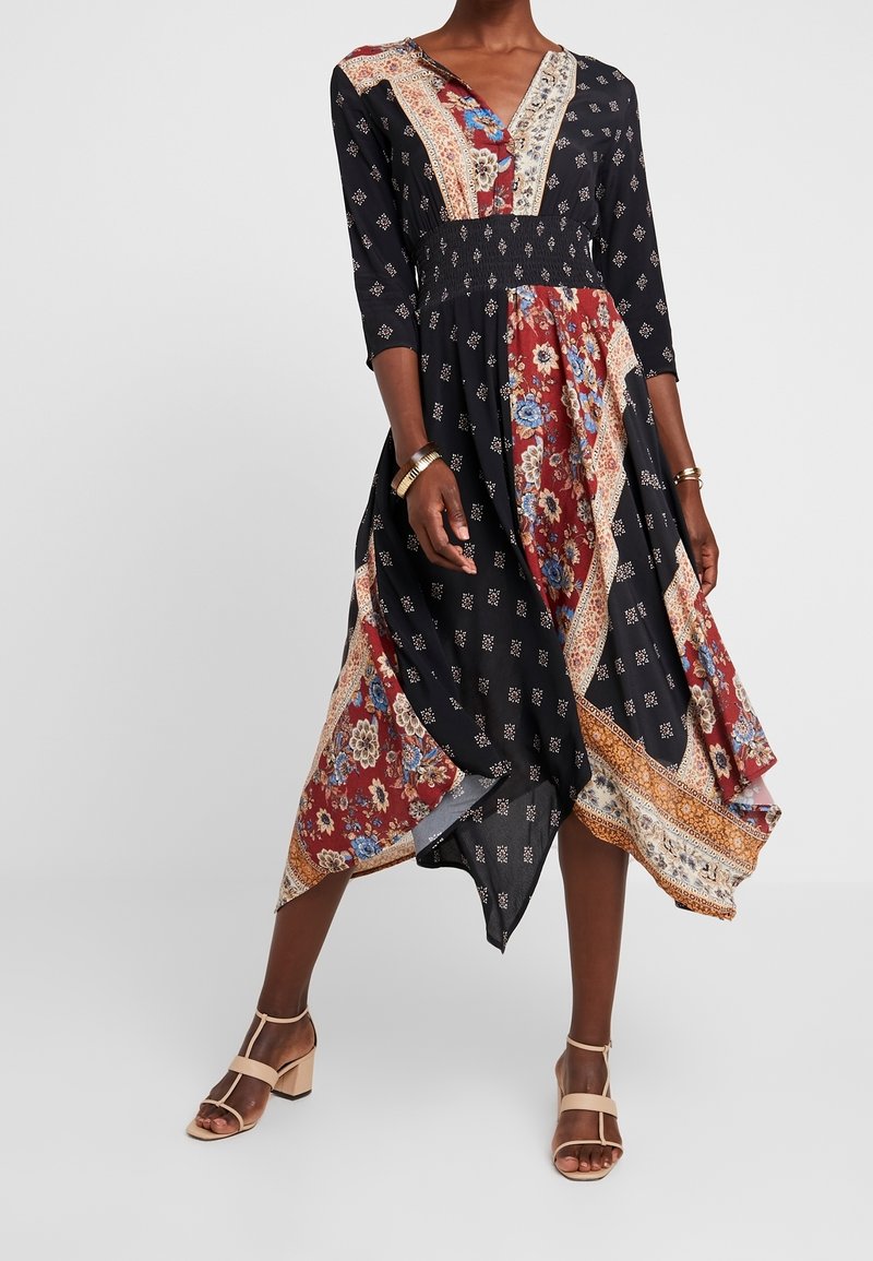 Femme portant une robe midi patchwork florale noire et rouge avec manches trois-quarts et sandales beiges à talons blocs.