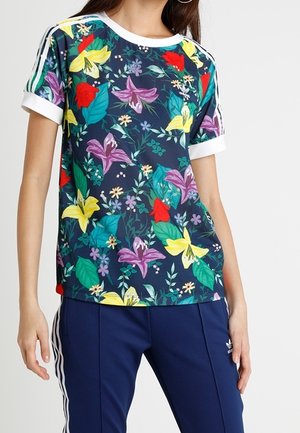 Femme portant un T-shirt bleu marine à fleurs jaunes, rouges, violettes avec une bordure blanche, associé à un pantalon de survêtement Adidas bleu foncé avec des bandes blanches.