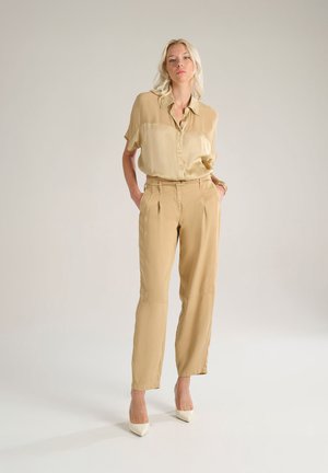 Camicia in seta beige con vestibilità rilassata e maniche corte, abbinata a pantaloni beige sartoriali e décolleté a punta di colore chiaro.