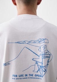 T-shirt branca com uma ilustração em azul de um urso a pescar, sentado numa cadeira, com montanhas ao fundo e texto abaixo.