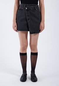 Zwarte denim mini-jurk met een wikkelontwerp, zichtbare stiksels en voorzakken, gecombineerd met zwarte kniehoge sokken en platte loafers.