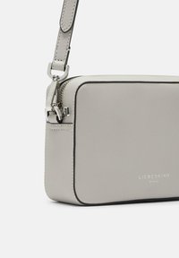 Bolso cruzado de cuero gris con forma rectangular, que cuenta con herrajes de plata, detalles de costura y un logo en la parte frontal.