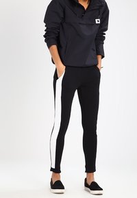 Black athletic leggings featuring a white side stripe, made of stretch fabric. Paired with a dark windbreaker and slip-on shoes.