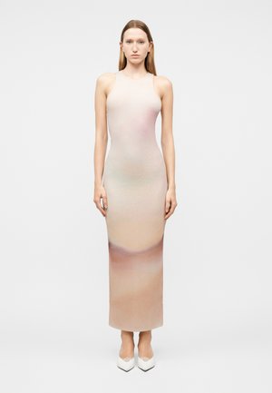 Ung kvinde med glat hår iført en ærmeløs, figursyet lang kjole med gradient fra beige til mauve og hvide spidse sko, stående stille.