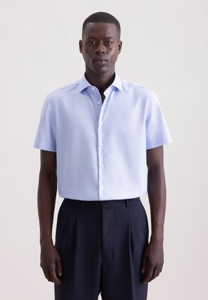 Mann trägt ein hellblaues, kurzärmliges Hemd mit Knöpfen, in hoch taillierte, dunkle, plissierte Hosen gesteckt, vor einem einfarbigen Hintergrund.