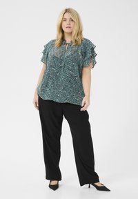 Blusa de color verde azulado, con estampado y mangas con volantes, que presenta un escote con lazo. Combinada con pantalones negros de pierna ancha. Zapatos negros de punta.