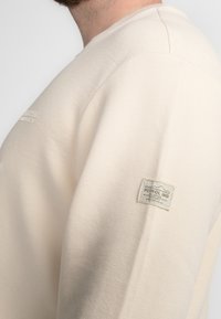 Crème kleurige trui met een gestructureerde stof, ronde halslijn en een klein logo-opdruk op de mouw. Naaddetails zichtbaar op de schouder.