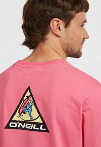 Camiseta de algodón rosa con un gráfico triangular negro y amarillo que presenta una figura de dibujos animados y el texto "O'NEILL" en la parte posterior. Diseño de cuello redondo.