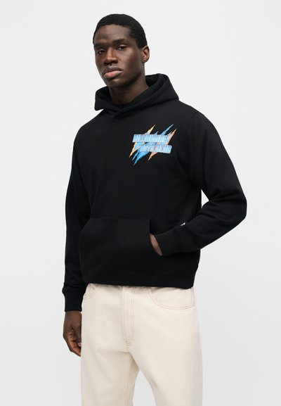 Νέος άνδρας που φοράει μαύρη μπλούζα με κουκούλα με μπλε και πορτοκαλί λογότυπο "Billionaire Boys Club", συνοδευόμενη από εκρού παντελόνι, στέκεται μπροστά σε απλό φόντο.