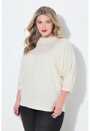 Kvinde med langt blondt hår iført en oversized hvid tekstureret sweater og sorte jeans, poserer foran en hvid baggrund.