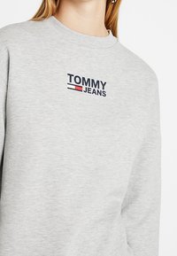 Light gray sweatshirt with "TOMMY JEANS" logo and red, white, and blue flag emblem on the chest, worn by a person.