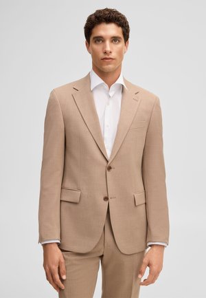 Mężczyzna z krótkimi kręconymi włosami, ubrany w beżowy garnitur na dwa guziki i białą koszulę, stojący na jednolitym jasnym tle.