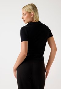 Čierny krátky rukáv top s priliehavým strihom a jemnou textúrou, zobrazený zozadu. Vykrojenie je nenápadné, dopĺňa elegantnú siluetu.
