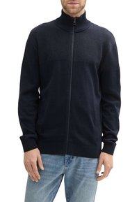 Granatowy sweter ze zamkiem błyskawicznym, z teksturowaną górną częścią, prążkowanymi mankietami i dołem. Posiada wysoki kołnierz oraz gładką dzianinę.