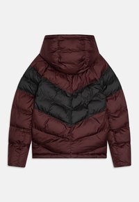 Maroon ja must puffer-jakk, millel on chevron-muster, kapuuts ja tikitud tekstuur. Kujundus rõhutab kontrastseid värve ja kujundeid.